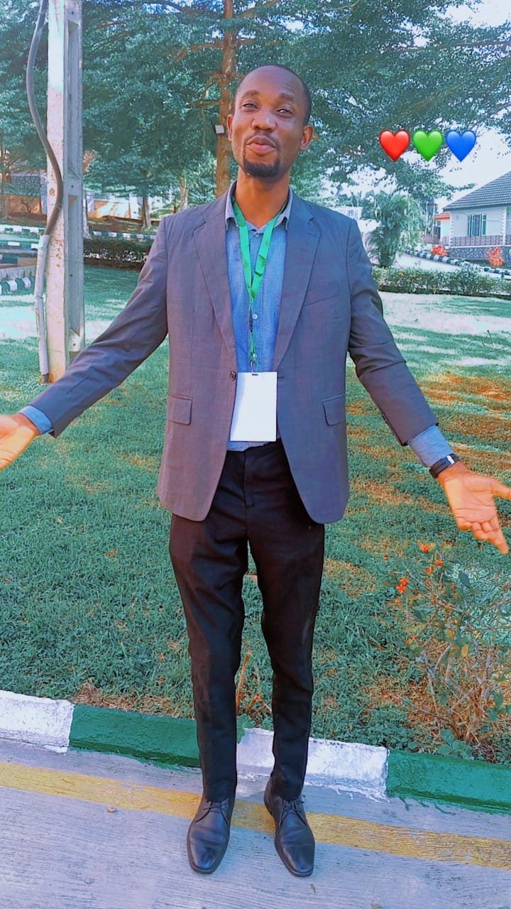 A man in a grey blazer and blue shirt stands outdoors on a lawn with his arms open, wearing a green lanyard.