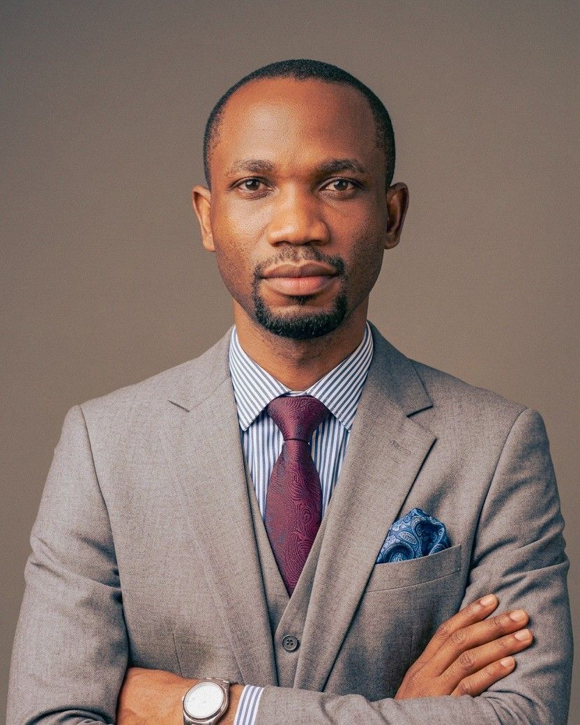 Tunde Godfrey in a light grey three-piece suit and burgundy tie, arms folded, looking at the camera